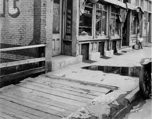In 1929, this section of 12th Street between Euclid and Garfield, showed one of the problems the city struggled with in trying to keep its sidewalks in good order: one property owner chose a wooden sidewalk, while the owner next door choose cement. Water standing on the street and decayed curbs also show that poorly maintained sidewalks are not a new problem. Courtesy Kansas City Public Library/ Missouri Valley Special Collections. 